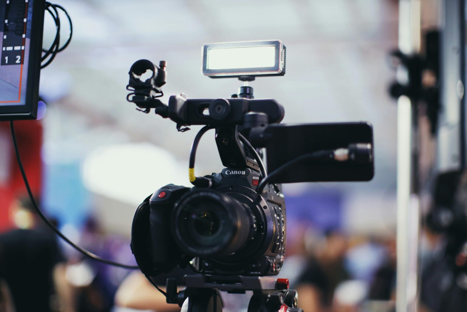 A close-up shot of a Canon video camera with attached equipment in a blurred indoor setting.